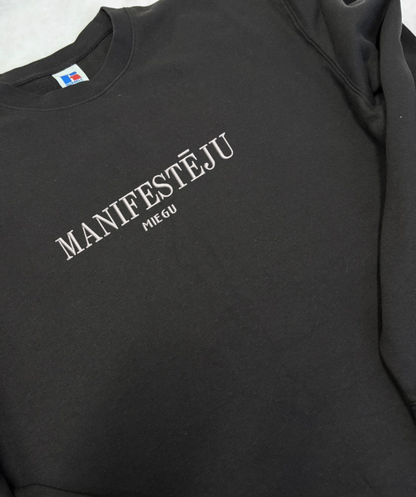 Izšūts teksts “manifestēju” uz melna unisex džempera